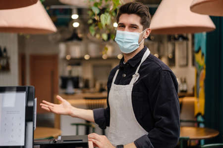 Young man in apron wearing protective medical mask standing at the cash register in coffee shop