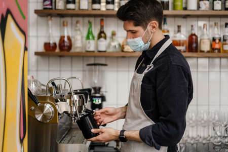 Young white brunette man in apron and protective mask working with coffee machineの写真素材