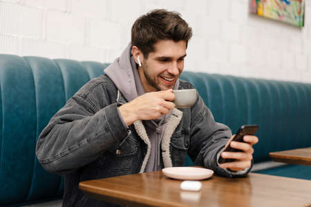 Smiling young casual man in earphones listening to music, holding mobile phone while sitting in cafe...