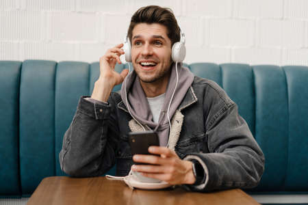 Smiling young casual man in headphones listening to music, holding mobile phone while sitting in...