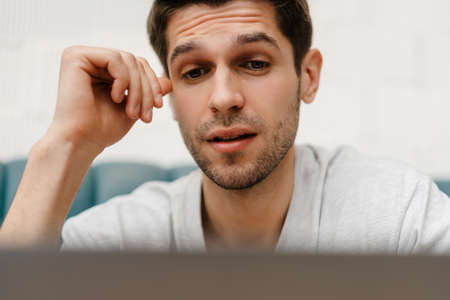 Confused young man student sitting in cafe indoors with laptop computer, looking at screenの写真素材