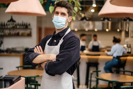 Young man in apron wearing protective medical mask standing at the cash register in coffee shopの写真素材