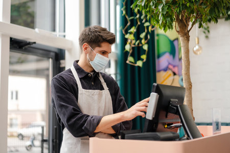 Young man in apron wearing protective medical mask standing at the cash register in coffee shopの写真素材