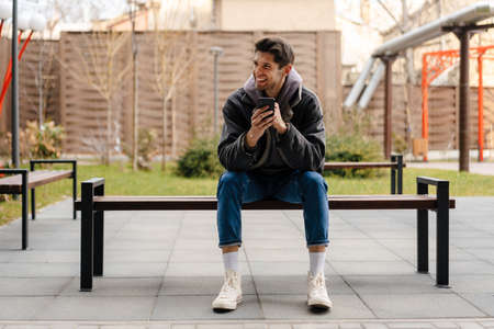 Smiling young white man in casual cothes using mobile phone outdoors in the city, sitting on a bench