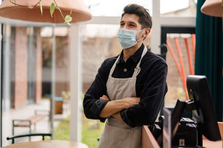Young man in apron wearing protective medical mask standing at the cash register in coffee shopの写真素材