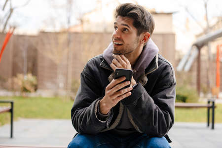 Smiling young white man in casual cothes using mobile phone outdoors in the city, sitting on a benchの写真素材