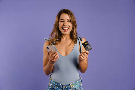 Young white woman holding credit card and cellphone isolated over purple background