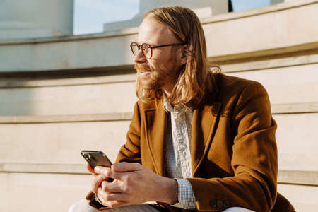 Ginger man smiling and using mobile phone while sitting on stairs outdoors