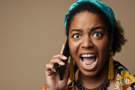 Young black woman screaming while talking on cellphone isolated over grey backgroundの写真素材