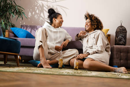 Two happy young african women friends sitting on a couch drinking beer eating popcorn at home...
