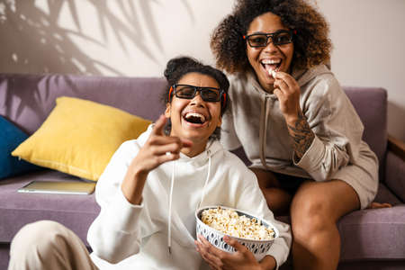 Two happy multiethnic women friends sitting on a couch at home with snacks watching TV laughingの写真素材