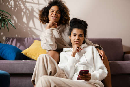 Two happy young african women sitting on a couch at home holding mobile phone looking at cameraの写真素材