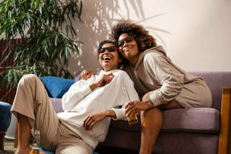 Two happy young african women friends sitting on a couch drinking beer at home watching TV in 3d...