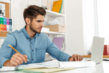 Young white man working with laptop and drawings in office indoors
