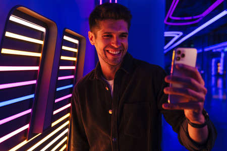 Young bristle man wearing shirt smiling while taking selfie on mobile phone indoors