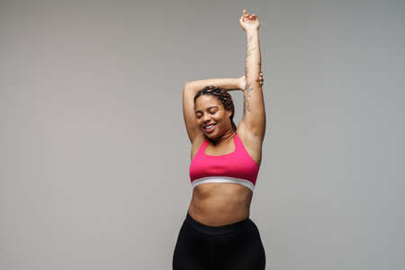 Smiling young african bodypositive fitness woman over gray wall background stretching exerciseの写真素材