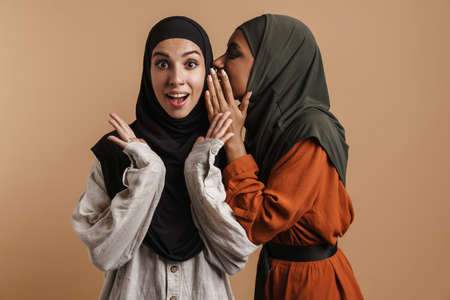 Young muslim woman in hijab whispering secret to her surprised friend isolated over beige backgroundの写真素材