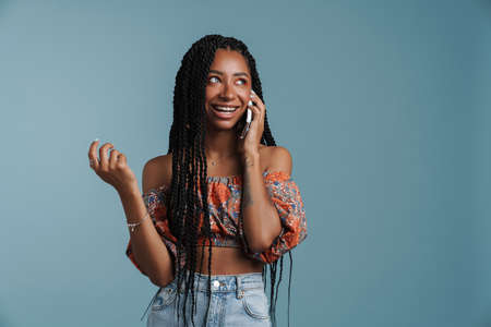 Happy african american young woman standing over blue background wearing summer look talking on...