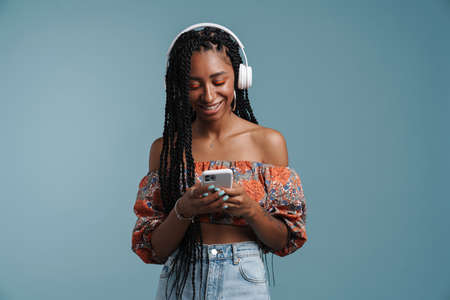 Happy african american young woman standing over blue background wearing summer look listening to...