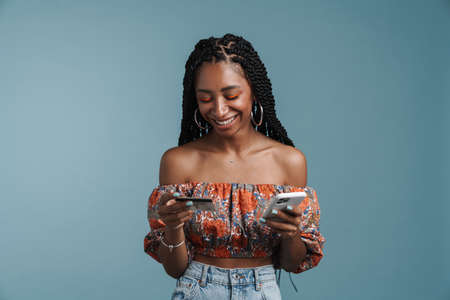 Happy african american young woman standing over blue background wearing summer look holding mobile...