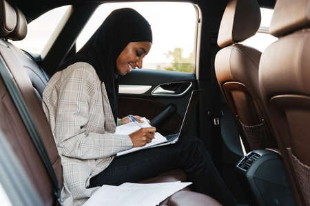 Black muslim woman wearing headscarf writing down notes while going in carの写真素材