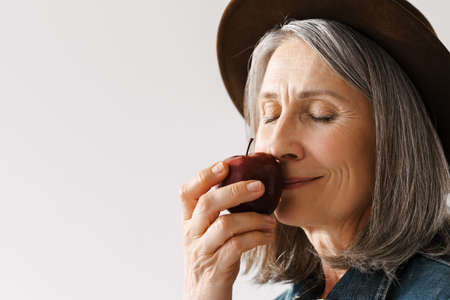 Grey senior woman in hat smiling while smelling apple isolated over white backgroundの写真素材