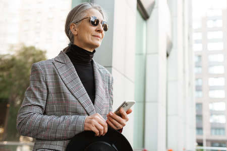 Grey white senior woman holding hat while using mobile phone using cellphone outdoorsの写真素材