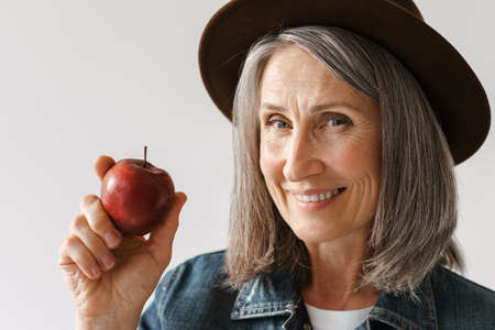 Grey senior woman in hat smiling while posing with apple isolated over white backgroundの写真素材