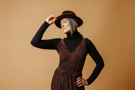 Grey senior woman wearing hat and dress posing at camera isolated over beige backgroundの写真素材