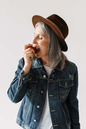 Grey senior woman in hat eating apple with her eyes closed isolated over white background