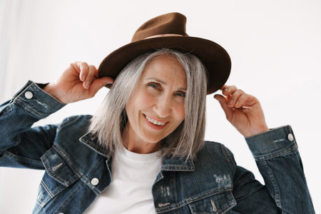 Grey senior woman smiling at camera while posing in hat isolated over white backgroundの写真素材