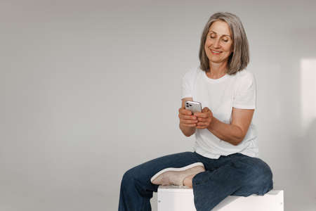 Grey senior woman using mobile phone while sitting on box isolated over white backgroundの写真素材