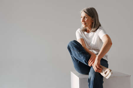 Grey senior woman in jeans smiling while sitting on box isolated over white backgroundの写真素材