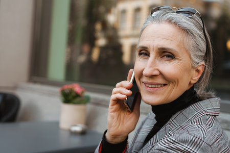 Grey white senior woman talking on cellphone while sitting in cafe outdoorsの写真素材