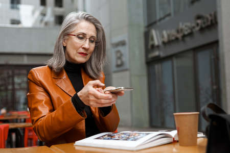 Grey white senior woman taking photo on cellphone while reading book in cafe outdoorsの写真素材