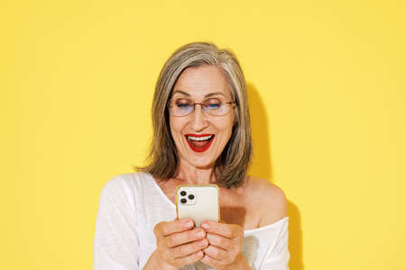 Mature woman in eyeglasses smiling while using mobile phone isolated over yellow wall