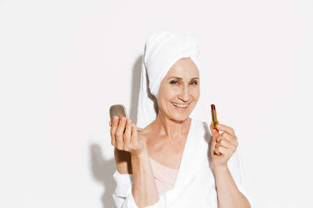 Senior woman wearing towel smiling while applying lipstick isolated over white wallの写真素材