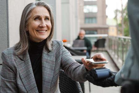 Grey white senior woman paying with mobile phone in cafe outdoorsの写真素材
