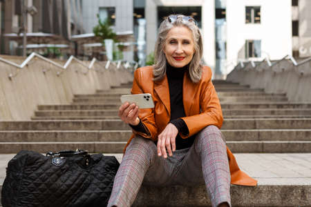 Grey white senior woman using mobile phone while sitting on stairs outdoorsの写真素材