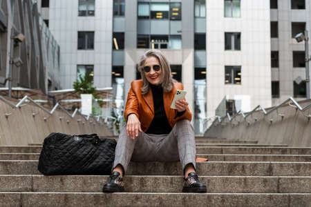 Grey white senior woman using mobile phone while sitting on stairs outdoors