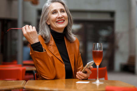Grey white senior woman using mobile phone while drinking wine in cafe outdoorsの写真素材