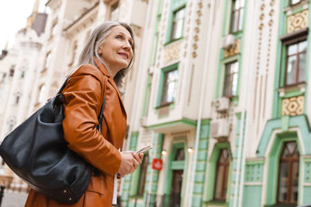 Grey senior woman using mobile phone while walking on city streetの写真素材