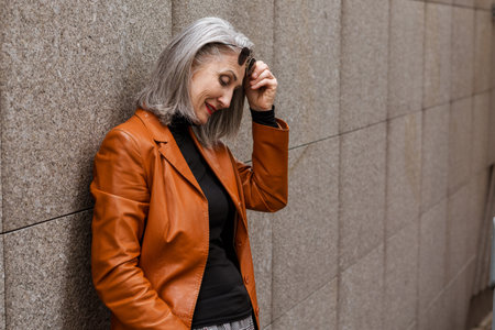 Grey senior woman in leather jacket smiling while leaning on wall outdoorsの写真素材
