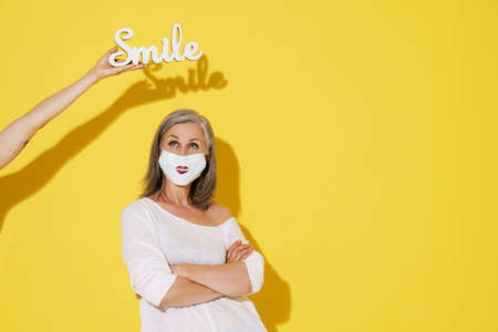 White senior woman in face mask posing with decorated white word isolated over yellow wallの写真素材