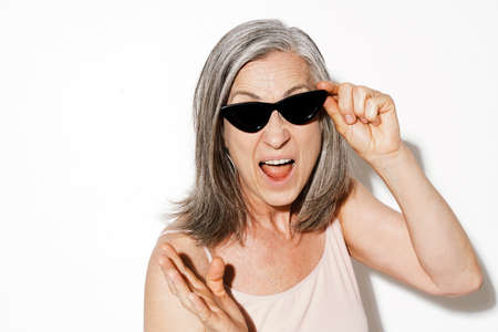 Mature woman in sunglasses gesturing while screaming at camera isolated over white wall