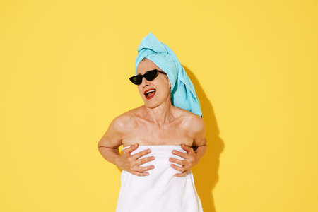 Mature woman in sunglasses holding her bust while making fun isolated over yellow wallの写真素材