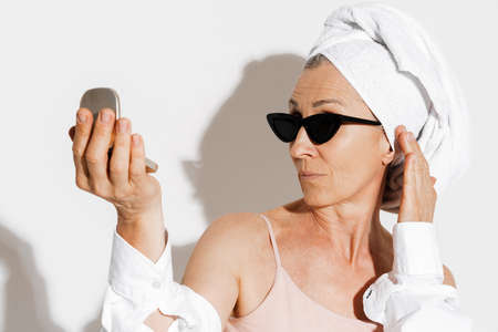Mature european woman in sunglasses looking at powder mirror isolated over white wallの写真素材