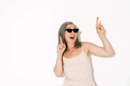 Senior woman wearing sunglasses laughing while pointing fingers upward isolated over white wallの写真素材