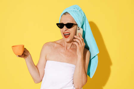 Senior woman wearing towels talking on cellphone while drinking coffee isolated over yellow wallの写真素材