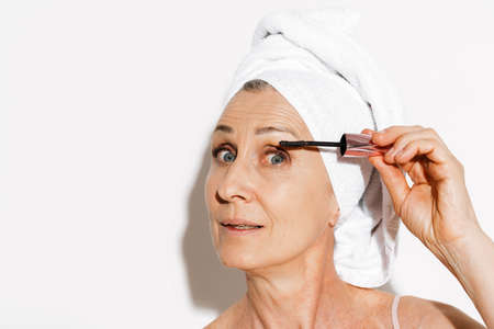 Senior woman wrapped in towel applying mascara while doing makeup isolated over white wallの写真素材